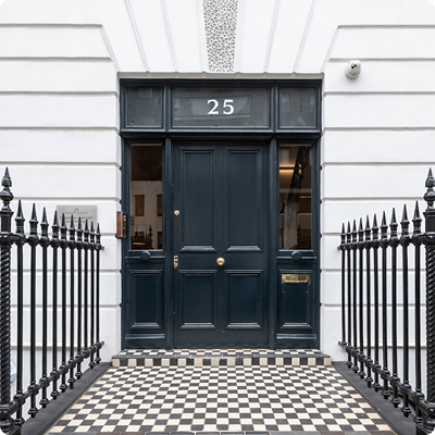 The entrance of a private clinic with a black door and metal fencing surrounded by white walls and a chequered tile floor